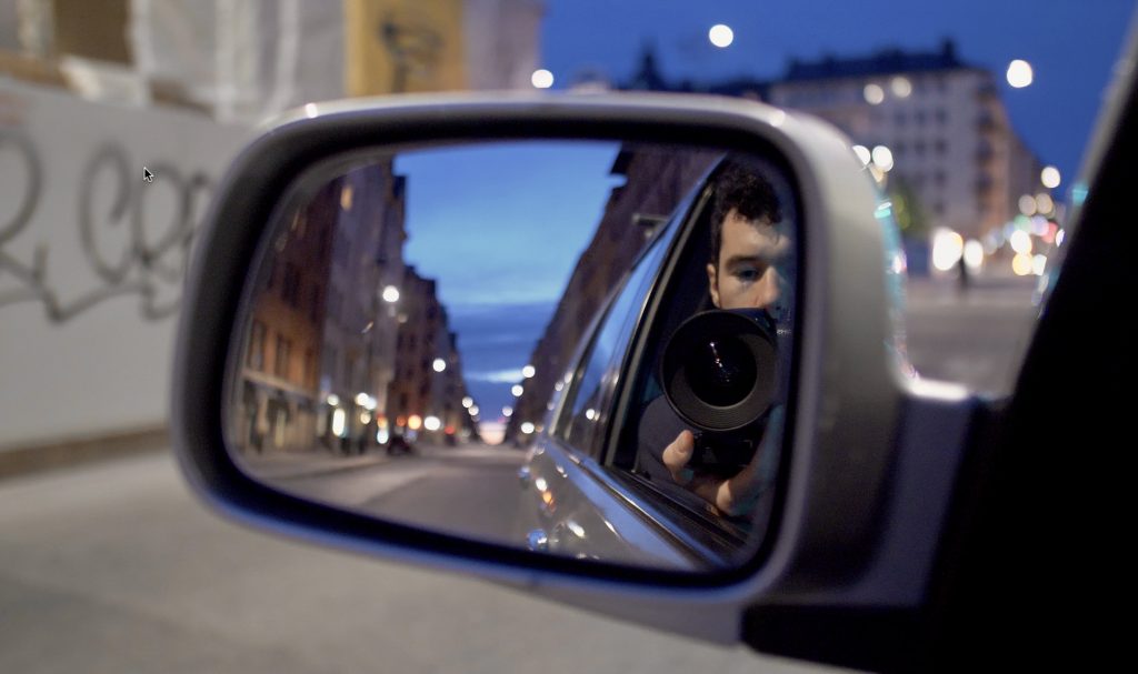 kursdeltagare på dokumentärfilmskolan som riktar en kameralins mot en backspegel Fotografi av en backspegel på en stad i skymning med en kursdeltagare på dokumentärfilmskolan som riktar en kameralins mot backspegeln.