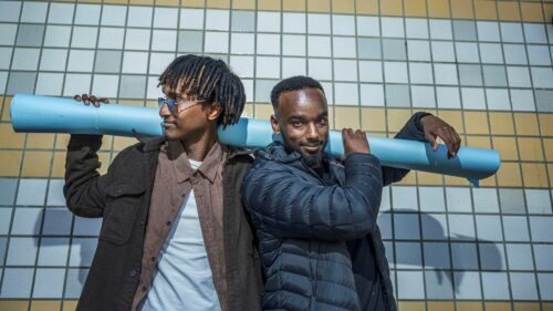 Två unga män står framför en rutig vägg och håller en ljusblå tub på axlarna.