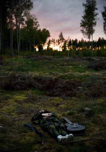 En ung man ligger på marken i skogen med en gitarr bredvid sig. Solen går ner i bakrgunden.