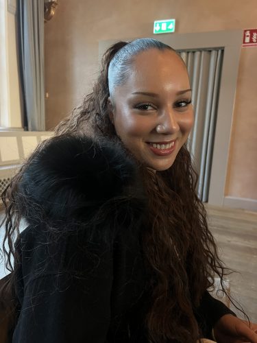 En ung kvinna med svart jacka och långt uppsatt hår ler mot kameran.